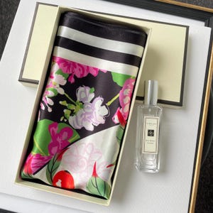 May include: A rectangular gift box containing a folded silk scarf with a black, white, and pink floral pattern and a black and white striped border, alongside a clear glass bottle of Jo Malone Red Roses cologne with a silver cap and white label.
