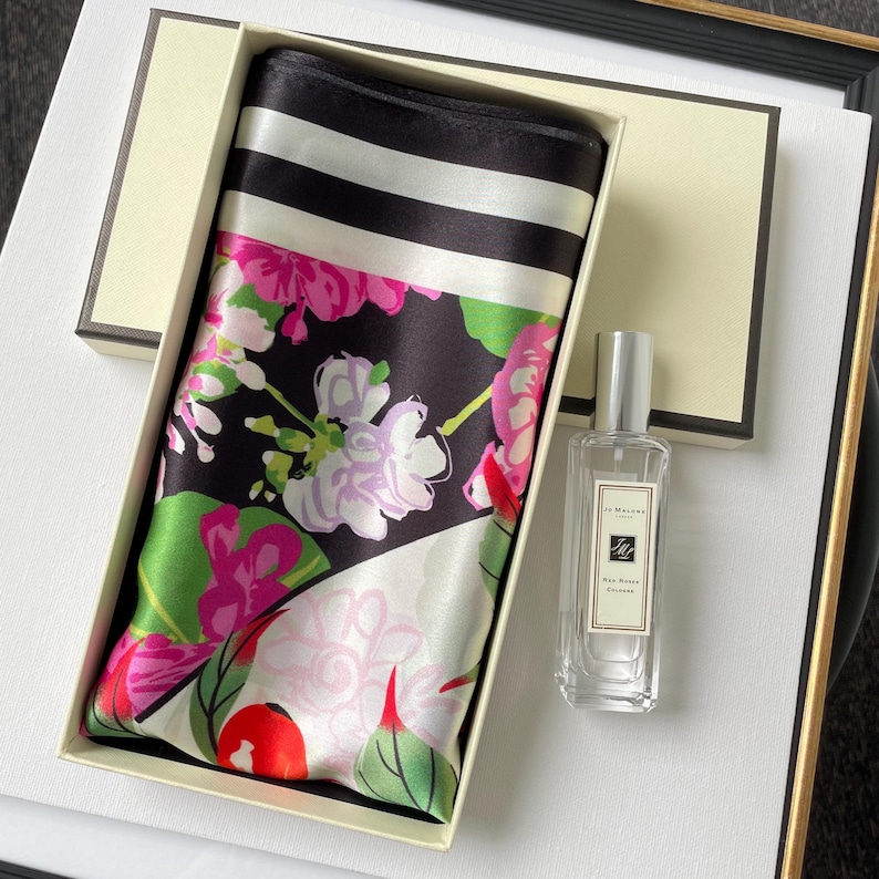 May include: A rectangular gift box containing a folded silk scarf with a black, white, and pink floral pattern and a black and white striped border, alongside a clear glass bottle of Jo Malone Red Roses cologne with a silver cap and white label.