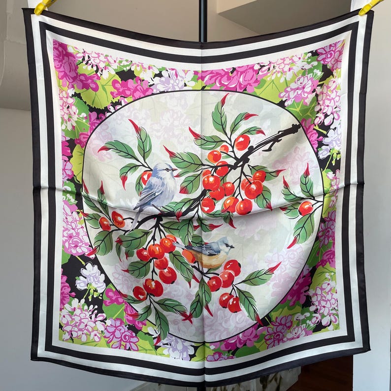 May include: A square scarf featuring a central circular design of two small birds perched on branches laden with bright red cherries and green leaves. The background displays clusters of pink and white flowers, framed by a bold black and white border.
