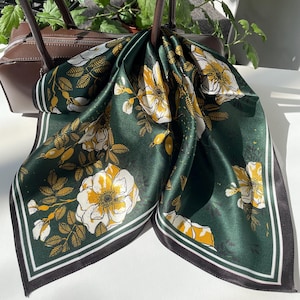 K&ouml;nnte beinhalten: Ein quadratisches Tuch mit einem tiefen waldgr&uuml;nen Hintergrund und einem sich wiederholenden Muster aus wei&szlig;en und gelben Wildrosenbl&uuml;ten, Knospen und Bl&auml;ttern. Das Tuch ist mit einem dunklen Rand und d&uuml;nnen wei&szlig;en Streifen an den Kanten versehen.