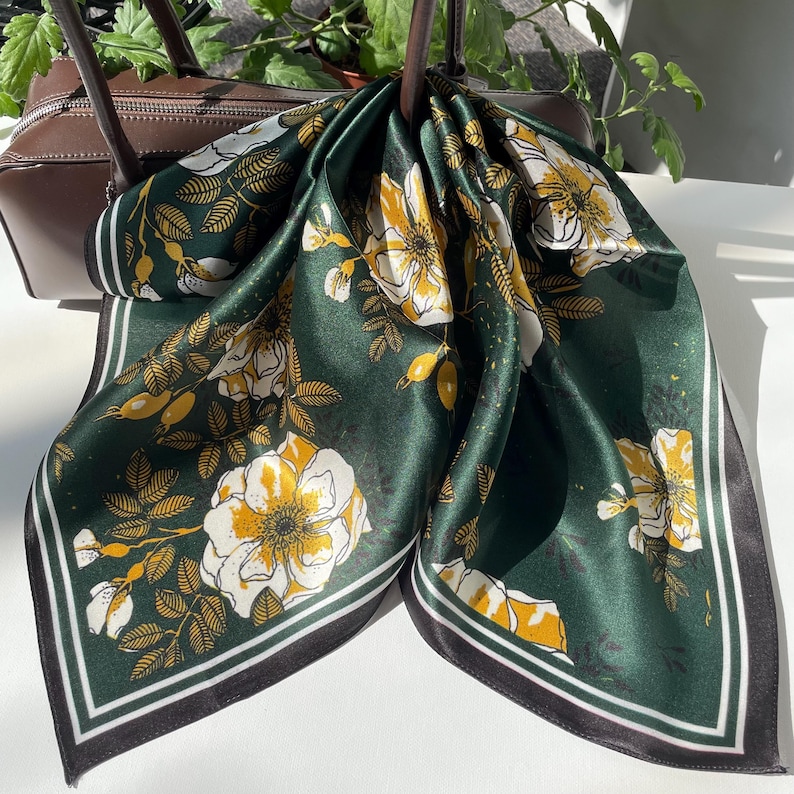 K&ouml;nnte beinhalten: Ein quadratisches Tuch mit einem tiefen waldgr&uuml;nen Hintergrund und einem sich wiederholenden Muster aus wei&szlig;en und gelben Wildrosenbl&uuml;ten, Knospen und Bl&auml;ttern. Das Tuch ist mit einem dunklen Rand und d&uuml;nnen wei&szlig;en Streifen an den Kanten versehen.