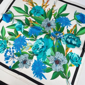 K&ouml;nnte beinhalten: Ein quadratisches Tuch mit wei&szlig;em Hintergrund, schwarzem Rand und einem lebendigen Blumenmuster. Das Design zeigt verschiedene blaue und t&uuml;rkisfarbene Bl&uuml;ten, gr&uuml;ne Bl&auml;tter und orangefarbene Akzente, die ein detailliertes botanisches Muster bilden.