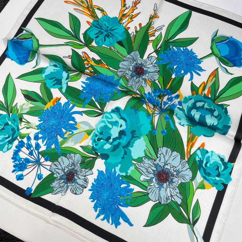 K&ouml;nnte beinhalten: Ein quadratisches Tuch mit wei&szlig;em Hintergrund, schwarzem Rand und einem lebendigen Blumenmuster. Das Design zeigt verschiedene blaue und t&uuml;rkisfarbene Bl&uuml;ten, gr&uuml;ne Bl&auml;tter und orangefarbene Akzente, die ein detailliertes botanisches Muster bilden.