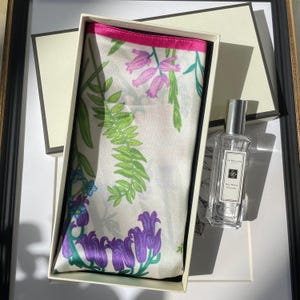 May include: A white silk scarf with a vibrant floral print featuring purple bell-shaped flowers, green fern leaves, and pink blossoms, finished with a bright pink border. Beside it sits a clear glass bottle of Jo Malone Red Roses cologne.