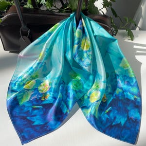 K&ouml;nnte beinhalten: Ein quadratisches Seidentuch mit einem lebendigen Farbverlauf in T&uuml;rkis, Cyan und Dunkelblau. Der Stoff ist mit einem blumigen Aquarellmuster in Gelb- und Hellgr&uuml;nt&ouml;nen verziert und &uuml;ber eine dunkelbraune Lederhandtasche drapiert.