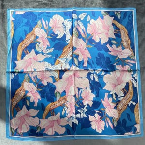 May include: A square scarf featuring a vibrant blue background with a repeating pattern of light pink flowers and tan, textured tree branches. The design includes a thin white border framing the floral and botanical illustration on the fabric.