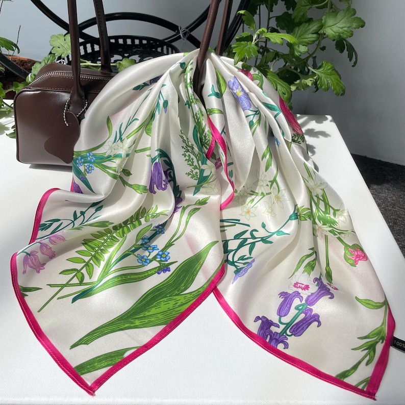 May include: A square silk scarf featuring a white background with a botanical print of green ferns, blue forget-me-nots, and purple bell-shaped flowers. The scarf has a vibrant hot pink border and is draped over a dark brown leather handbag.