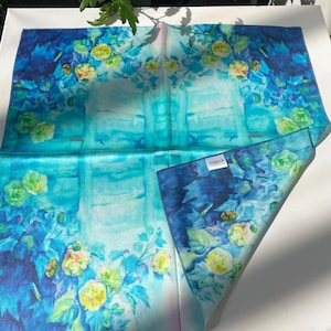 K&ouml;nnte beinhalten: Ein quadratisches Seidentuch mit einem lebendigen Blumenmuster in den Farben Dunkelblau, T&uuml;rkis und leuchtendem Cyan. Das Design zeigt Ansammlungen von gelben und hellgr&uuml;nen Bl&uuml;ten mit gr&uuml;nen Bl&auml;ttern sowie ein kleines, wei&szlig;es, rechteckiges Etikett.