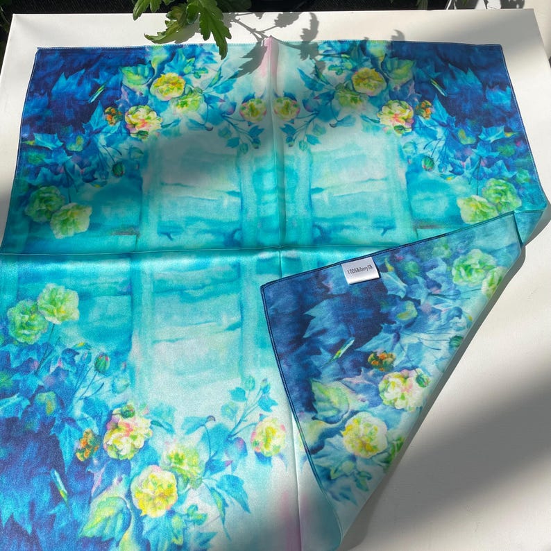 K&ouml;nnte beinhalten: Ein quadratisches Seidentuch mit einem lebendigen Blumenmuster in den Farben Dunkelblau, T&uuml;rkis und leuchtendem Cyan. Das Design zeigt Ansammlungen von gelben und hellgr&uuml;nen Bl&uuml;ten mit gr&uuml;nen Bl&auml;ttern sowie ein kleines, wei&szlig;es, rechteckiges Etikett.