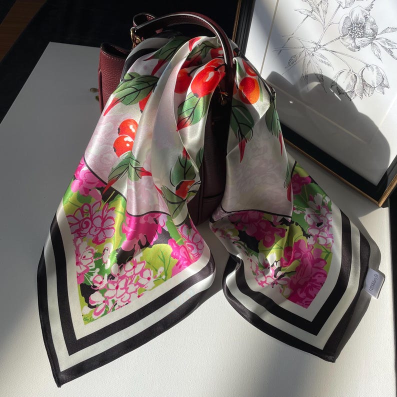 May include: A square silk scarf featuring a vibrant pattern of red cherries, green leaves, and bright pink flowers on a white background, finished with a black and white striped border. The scarf is draped over a dark burgundy leather handbag.