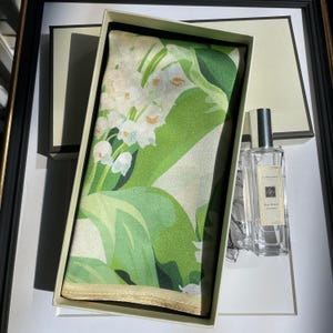 K&ouml;nnte beinhalten: Ein rechteckiges Seidentuch mit einem wei&szlig;en Maigl&ouml;ckchen-Blumenmuster auf gr&uuml;nem Hintergrund, pr&auml;sentiert in einer cremefarbenen Schachtel neben einem durchsichtigen Glasflakon Jo Malone Red Roses Cologne mit silbernem Verschluss und wei&szlig;em Etikett.