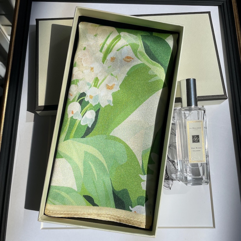 K&ouml;nnte beinhalten: Ein rechteckiges Seidentuch mit einem wei&szlig;en Maigl&ouml;ckchen-Blumenmuster auf gr&uuml;nem Hintergrund, pr&auml;sentiert in einer cremefarbenen Schachtel neben einem durchsichtigen Glasflakon Jo Malone Red Roses Cologne mit silbernem Verschluss und wei&szlig;em Etikett.