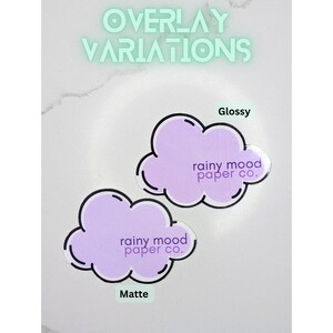 May include: Two purple cloud-shaped stickers with the text "rainy mood paper co." in black text. The sticker on the left has a matte finish and the sticker on the right has a glossy finish.