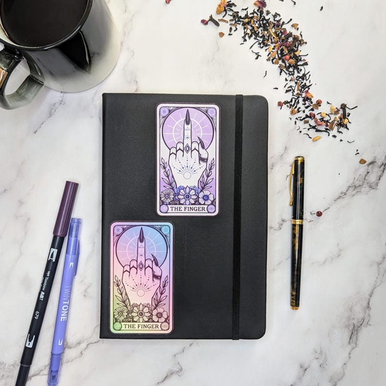 May include: Two holographic stickers with a middle finger design and the text "The Finger" on a black notebook. The stickers have a purple and blue gradient.