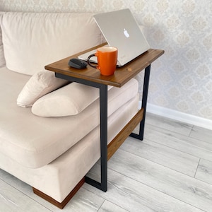 May include: A wooden side table with a dark brown top and black metal legs. An open laptop, a black computer mouse, and an orange mug are on the table. The table is next to a beige sofa.