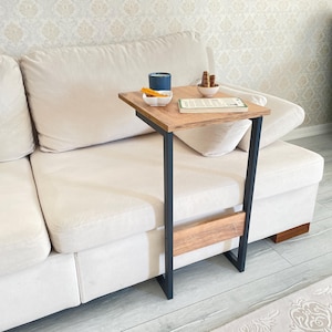 Pode incluir: Uma mesa de apoio de madeira com uma estrutura de metal preto, presa a um sofá branco. A mesa tem uma prateleira de madeira marrom e um tampo retangular de madeira.