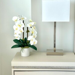 May include: A white orchid arrangement in a white pot, a silver lamp with a white shade, and a cream-colored nightstand with a crystal-like drawer pull. The orchid has white petals with yellow centers and green leaves.