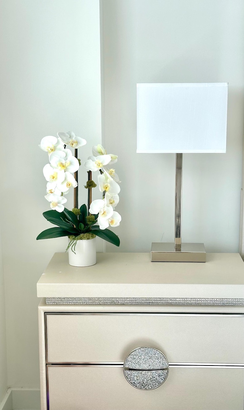 May include: A white orchid arrangement in a white pot, a silver lamp with a white shade, and a cream-colored nightstand with a crystal-like drawer pull. The orchid has white petals with yellow centers and green leaves.