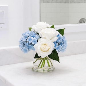 May include: A floral arrangement in a clear glass vase. The arrangement features white roses and light blue hydrangeas with green leaves. The vase sits on a white countertop, with a mirror in the background.