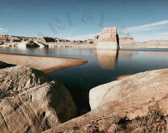May include: A large, tan rock formation rises from a blue lake. The water is calm and reflects the sky and the rock. The shore is sandy and there are smaller rock formations in the background.