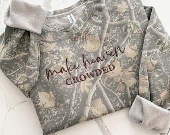 May include: A long-sleeve sweatshirt featuring a camouflage pattern of green, brown, and tan leaves and branches. The text "make heaven CROWDED" is embroidered in dark brown thread across the center chest. The interior is lined with gray fleece.