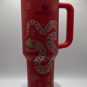May include: Red Stanley tumbler with a silver snake design and the Stanley logo. The tumbler has a handle and a clear lid. Decorative Chinese characters and symbols are etched on the tumbler.