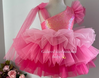May include: A pink tulle dress with a sequined bodice and a pink bow headband. The dress has multiple layers of tulle and is decorated with a pink bow. The dress is accompanied by a pair of pink shoes with a pink bow.