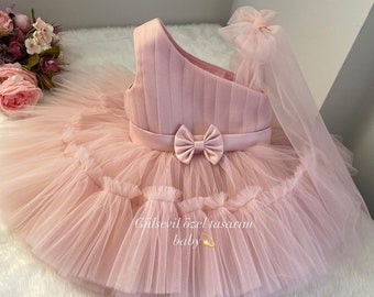 May include: A pink tulle baby dress with a satin bodice and a matching pink bow. The dress has a tulle veil attached to the shoulder strap. The dress is paired with a pink tulle headband with a satin bow and a pair of pink leather baby shoes with a sparkly bow.