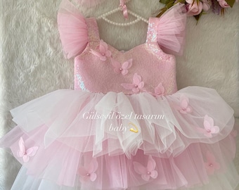 May include: A pink tulle baby dress with butterfly embellishments and a sequined bodice. The dress has a layered skirt with a white tulle bottom layer. The dress is on a white surface with a pink bow headband and a pearl necklace.