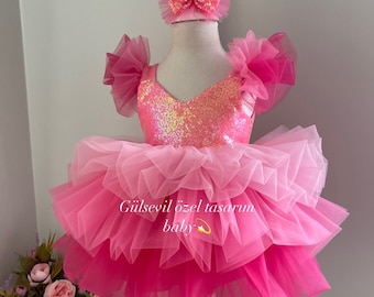 May include: A pink tulle dress with a sequined bodice and ruffled sleeves. The dress has a layered tulle skirt with a gradient of pink shades. The dress is on a white mannequin and has a pink bow headband. The text "Gülsevil özel tasarım baby" is on the dress.