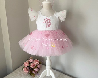May include: A pink tulle tutu dress with a white bodice featuring a pink sparkly letter "B" and pink heart accents. The dress is on a white mannequin with a pink bow headband. The text "Gülsevil özel tasarım baby" is visible on the dress.