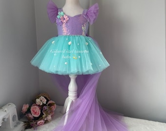 Op de afbeelding: Een lichtpaarse en turquoise tulejurk met een high-low zoom. De jurk heeft een paillettenlijfje met een bloemversiering en een parelrand. De jurk staat op een witte pop en heeft een bijpassende lichtpaarse tule sleep. De tekst "Gülsevil özel tasarım baby" is op de jurk gedrukt.