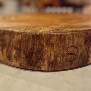 May include: A large, round wooden cutting board with a natural wood grain finish. The board has a dark brown color and a slightly uneven surface. The board is made from a single piece of wood and has a natural edge. The board has a small, circular logo burned into the side.