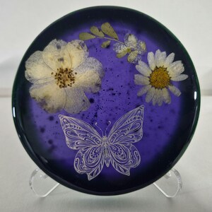 May include: A circular, deep purple resin coaster featuring embedded pressed white flowers and a delicate, white line-art butterfly illustration. The surface has a smooth, glossy finish with subtle dark speckles throughout the translucent material.