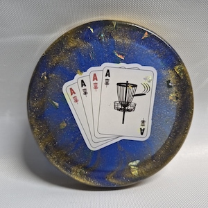 May include: A round coaster with a blue and gold resin design. The coaster features four playing cards with the Ace of each suit, and a disc golf basket graphic. The coaster is a unique accessory.