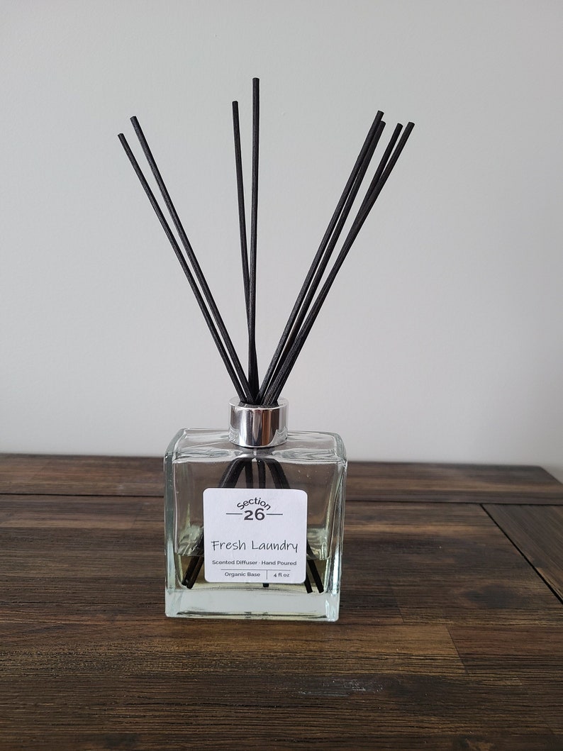 Puede incluir: Una botella de vidrio transparente con tapa plateada y varillas difusoras negras. La botella contiene un l&iacute;quido amarillo claro y tiene una etiqueta que dice "Section 26 Fresh Laundry Scented Diffuser - Hand Poured Organic Base 4 fl oz".