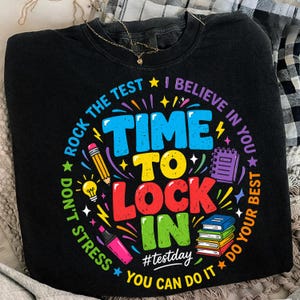 Könnte beinhalten: Schwarzes Sweatshirt mit einem farbenfrohen Aufdruck, der "Time to Lock In" zeigt, zusammen mit Bildern von Schulmaterialien. Der Text enthält "Rock the Test", "I Believe in You" und "You Can Do It".