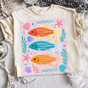Pu&ograve; includere: T-shirt color crema con una grafica colorata di tre pesci a strisce in rosso, blu e arancione. Il design include conchiglie, stelle marine e piante acquatiche su uno sfondo a righe rosa. Un design divertente e stravagante.