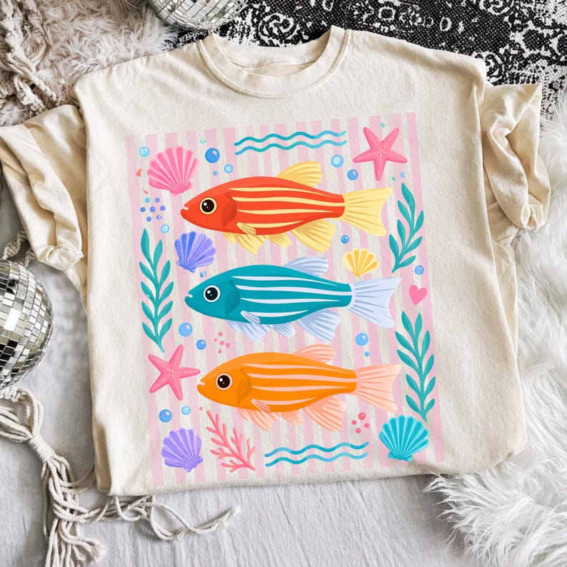 Pu&ograve; includere: T-shirt color crema con una grafica colorata di tre pesci a strisce in rosso, blu e arancione. Il design include conchiglie, stelle marine e piante acquatiche su uno sfondo a righe rosa. Un design divertente e stravagante.