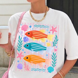 Pu&ograve; includere: T-shirt bianco con un colorato design acquatico. La grafica include tre pesci a strisce arancioni, blu e gialli, circondati da conchiglie, stelle marine e alghe. Lo sfondo presenta strisce rosa e bianche.