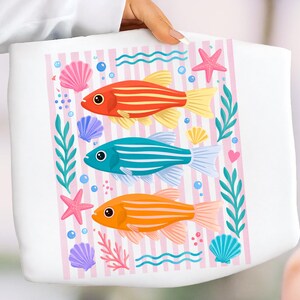Pu&ograve; includere: Borsa tote bianca con un disegno di tre pesci a strisce rossi, blu e arancioni. La borsa presenta uno sfondo a righe rosa e bianche, conchiglie, stelle marine e piante acquatiche. Il design ha un tema marino.