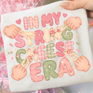 May include: A light gray t-shirt with the phrase "IN MY STRING CHEESE ERA" in pink, green, and yellow patchwork letters. The design includes floral and gingham patterns, with hands pulling string cheese.