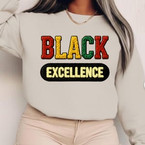 Può includere: Una felpa color crema con la scritta ricamata "BLACK EXCELLENCE" in rosso, giallo e verde. Sotto, un ovale nero mostra la parola "EXCELLENCE" ricamata in giallo.