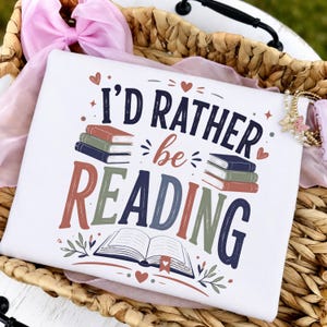 May include: A white t-shirt featuring the text I'D RATHER be READING in dark blue, red, and green lettering. The design includes illustrations of stacked books, an open book with a bookmark, small hearts, and decorative leaf accents.