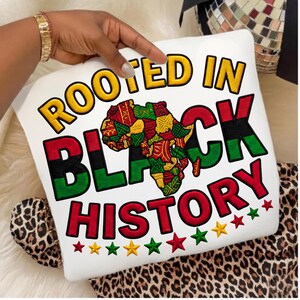 May include: White pillow with the text "ROOTED IN BLACK HISTORY" in yellow, red, and green. The design includes a colorful map of Africa and stars. The pillow is on a leopard print surface.