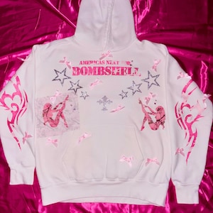 May include: White hoodie with pink and silver embellishments. The hoodie has the text "America's Next Top Bombshell" printed on the front. The hoodie also has pink bows and stars.