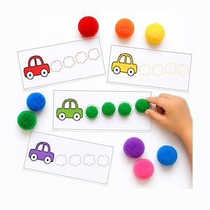 May include: Educational activity with toy cars and pom-poms. The image shows printed cards with car illustrations and cloud outlines, alongside red, orange, yellow, green, blue, purple, and pink pom-poms. A hand is placing a green pom-pom.