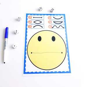 May include: A yellow smiley face game with a blue border, featuring numbered line variations. A blue marker and several dice are scattered around the game board. The game is designed for educational purposes.