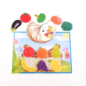 May include: A colourful set of felt fruit and vegetable shapes, including a banana, apple, pear, grapes, lemon, orange, aubergine, tomato, pumpkin, broccoli, and a green pepper. The shapes are arranged around a brown felt basket with a white felt bunny face on top. The bunny face has a pink nose and cheeks, and a blue bow.