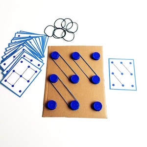 May include: A handmade educational toy featuring a cardboard base with blue bottle caps and rubber bands. Includes a set of blue-bordered cards with dot patterns and a collection of black rubber bands. The toy is designed for learning and play.